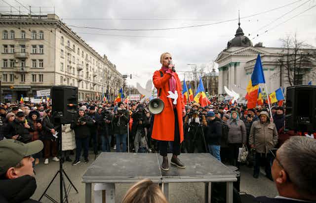 A woman with a loudhailer stands on a platform addressing a crowd waving Moldovan flags.