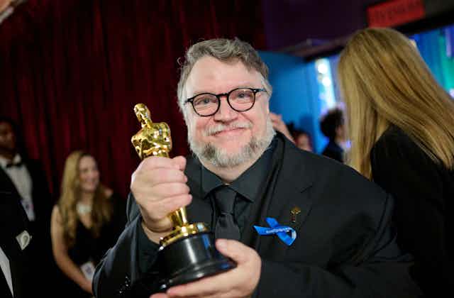 Guillermo del Toro poses backstage with his Oscar. He wears glasses and an all black suit.