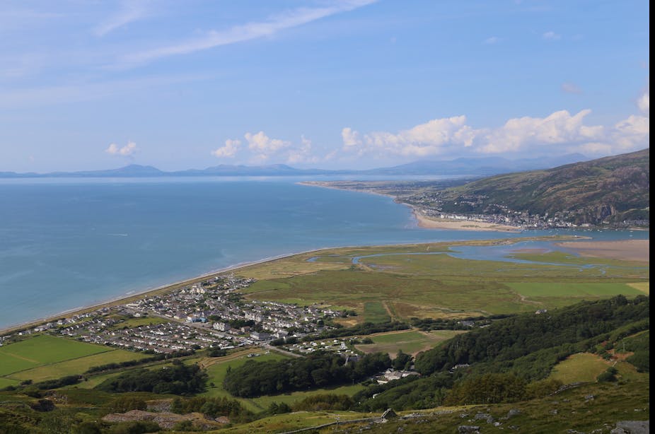 A view of a small village from above. It lies right next to the sea. There is a large, sandy estuary to the right of the village and a range of mountains in the distance. 