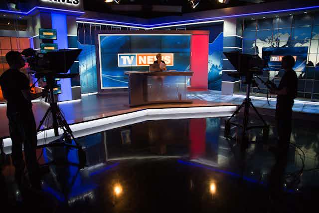 A TV newscast, with a woman anchor at a desk, being filmed by two cameras.