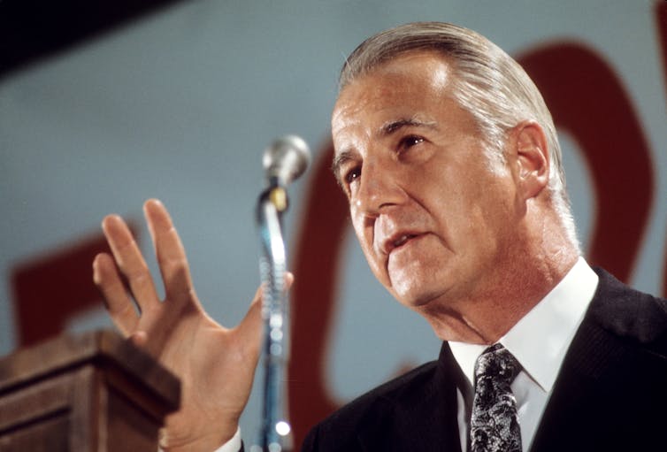 A man with a receding hairline and gray hair talking into a microphone.