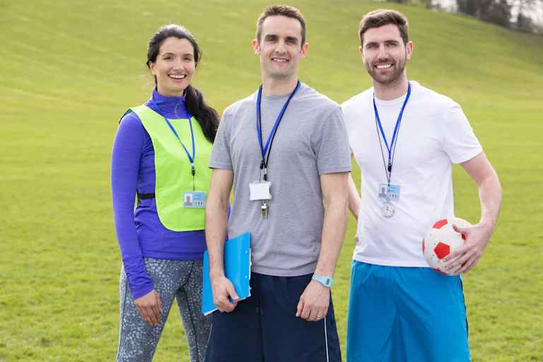 Una mujer y dos hombres sonrientes, con ropa deportiva y silbato y acreditación colgados del cuello.