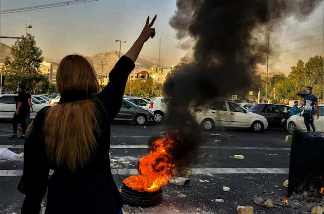 A long-haired woman is seen from behind giving the victory sign as people protests. A flaming tire is in front of her.