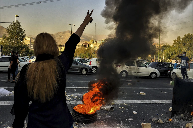 A long-haired woman is seen from behind giving the victory sign as people protests. A flaming tire is in front of her.