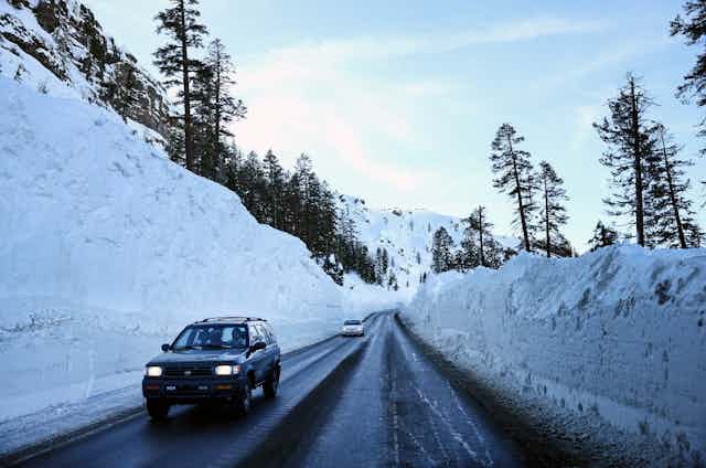 Cars drives through a plowed highway with snow more than twice their height on either side.