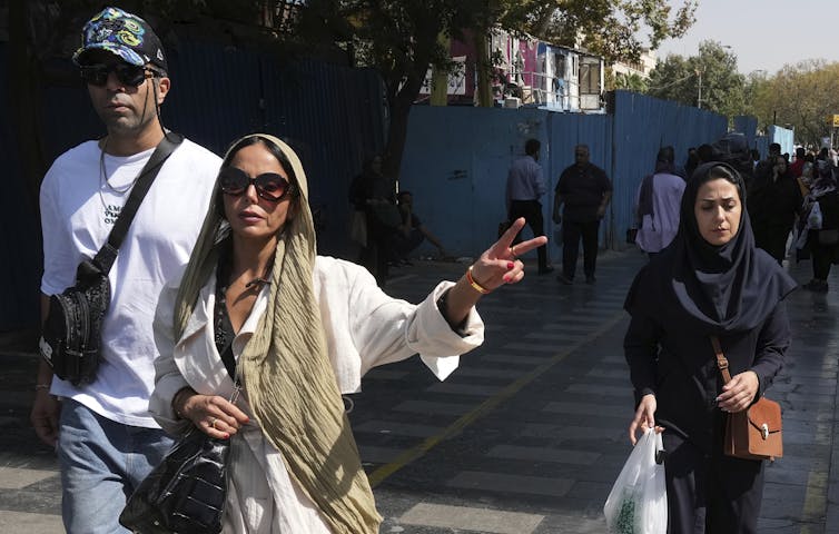A woman in white with a beige headscarf and dark sunglasses flashes a victory sign as she walks along a a street.