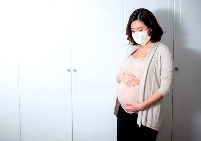 A pregnant woman wearing a face mask holds her stomach.