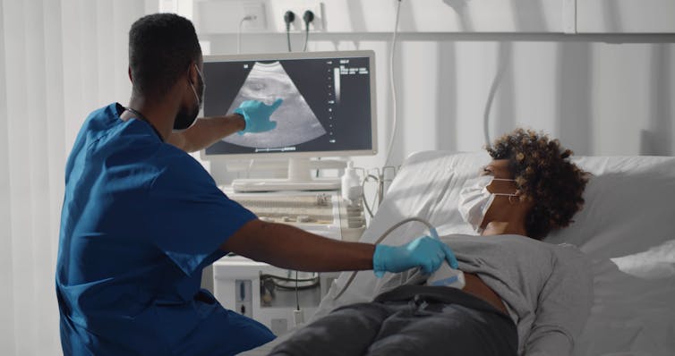 A woman lying on a clinic bed getting an abdominal ultrasound while a technician in scrubs points to an image on a screen