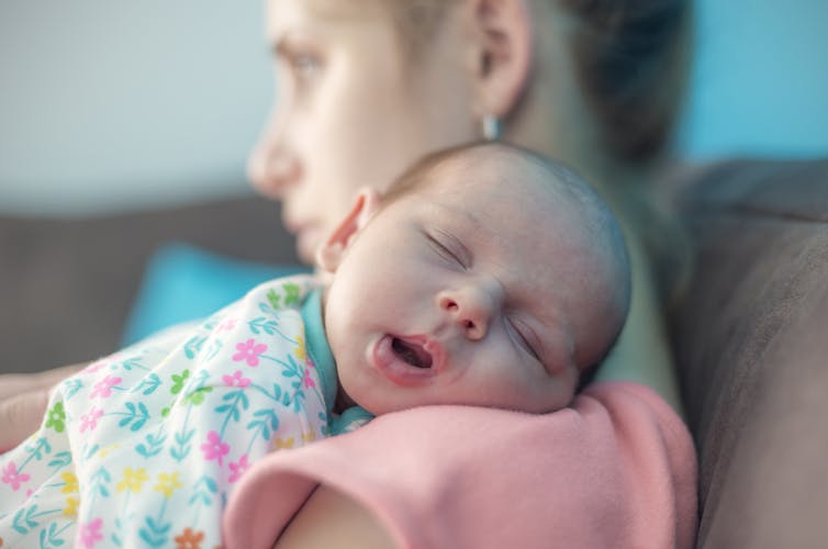 A woman slightly out of focus in the background with an infant asleep on her shoulder in the foreground