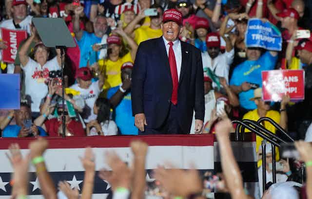 A man in a red Make American Great Again hat, blue suit and red die smiles and shouts to a crowd whose hands are raised towards him.
