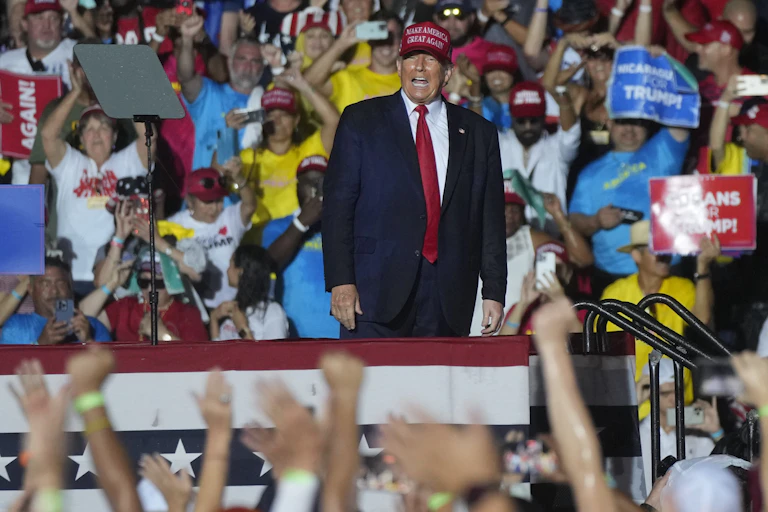 A man in a red Make American Great Again hat, blue suit and red die smiles and shouts to a crowd whose hands are raised towards him.