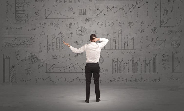 Man standing in front of grey wall full with graph pie charts and calculations, finance, economics, uncertain, confused, frustrated.