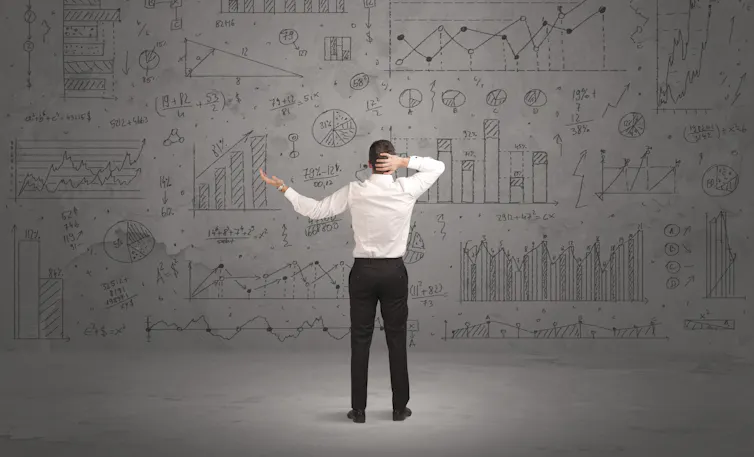 Man standing in front of grey wall full with graph pie charts and calculations, finance, economics, uncertain, confused, frustrated.