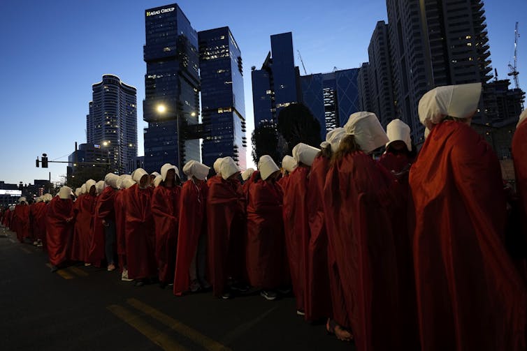 A row of women dressed in red robes and white bonnets stand in a row in front of skyscrapers at dusk.