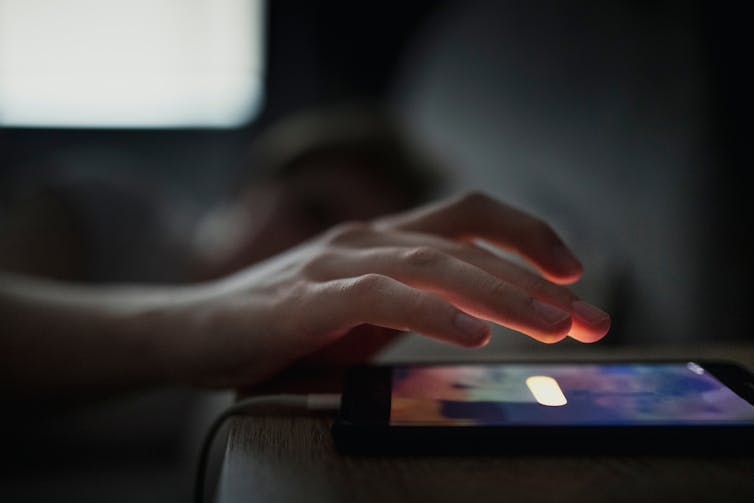 A hand of a sleeping person hovering over the alarm button on a phone next to their bed