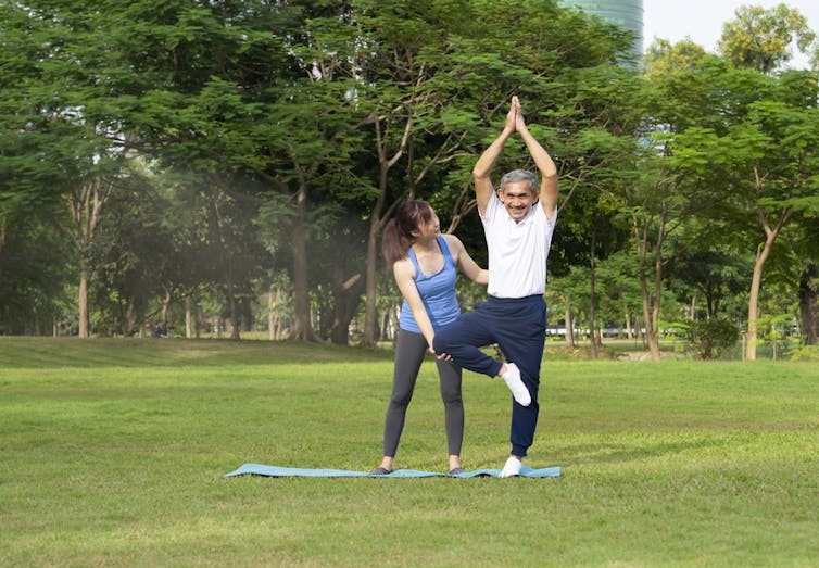 An older man standing on one leg in a park doing yoga while a younger woman in athletic wear guides him
