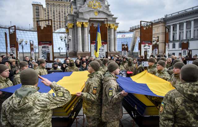A crowd of men with Ukrainian army uniforms salutes flag covered coffins in front of a church.