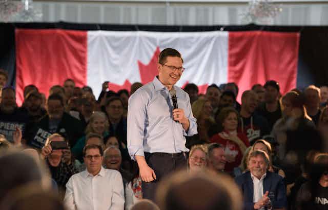 A dark-haired man in glasses smiles as he stands on a stage among a room of people.