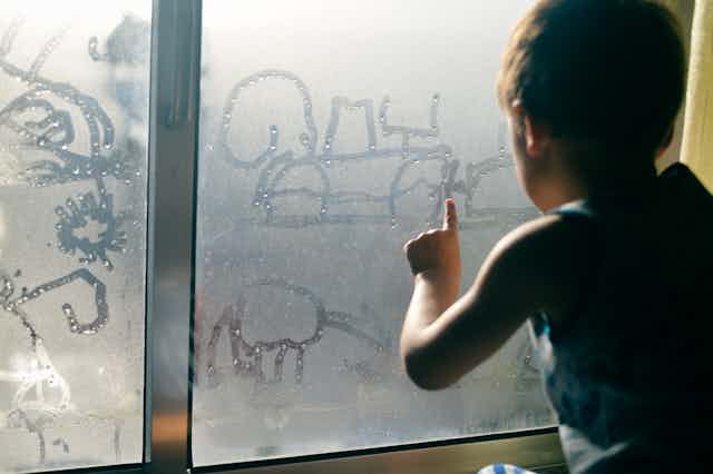 A young boy drawing in condensation on a window.