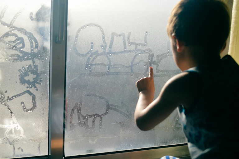A young boy drawing in condensation on a window.