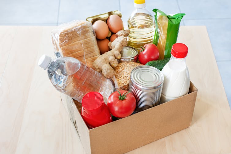 Cardboard box full of food including eggs, vegetables, bottled milk, oil, bread, and ginger root