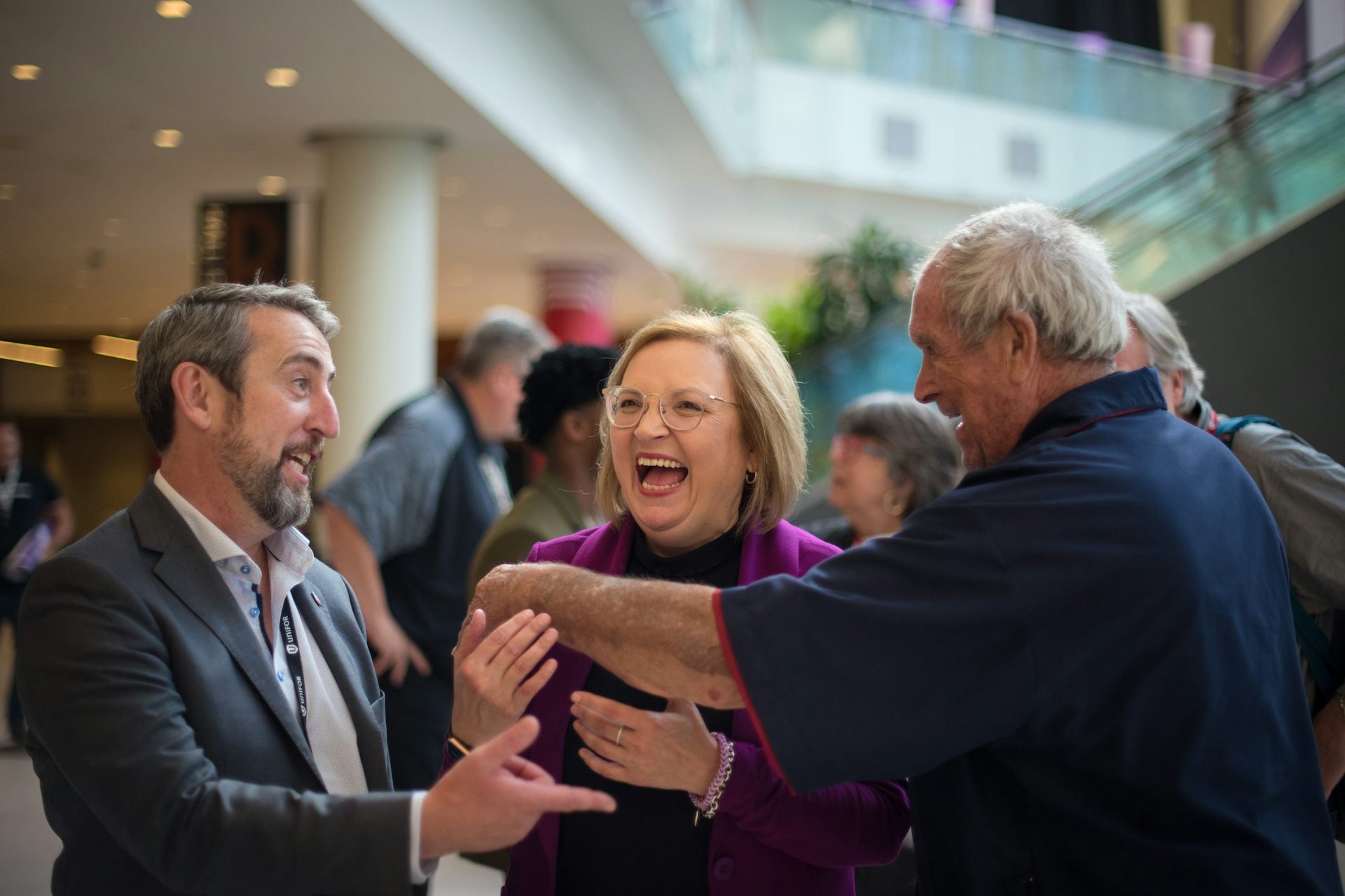A woman with dark blonde hair and glasses laughs as people surround her.