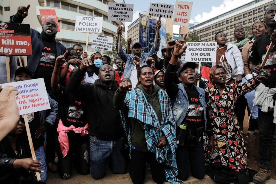 Men and women on their knees, fists held up in protest and holding up signs that say 'don't kill me, employ me' 'stop police impunity' and 'stop mass genocide on our youth'