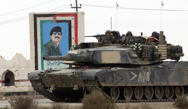 A dark tank has two soldiers sitting on top of it, peaking out, while it goes past a mural of a man with a black hat and dark glasses on a grey day. 