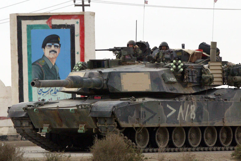A dark tank has two soldiers sitting on top of it, peaking out, while it goes past a mural of a man with a black hat and dark glasses on a grey day.