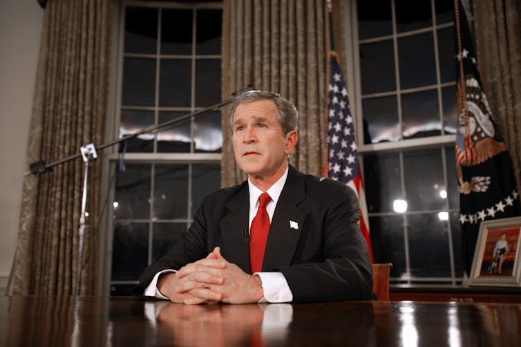 Un hombre blanco de pelo canoso viste traje oscuro y corbata roja y está sentado a una mesa con una bandera estadounidense detrás