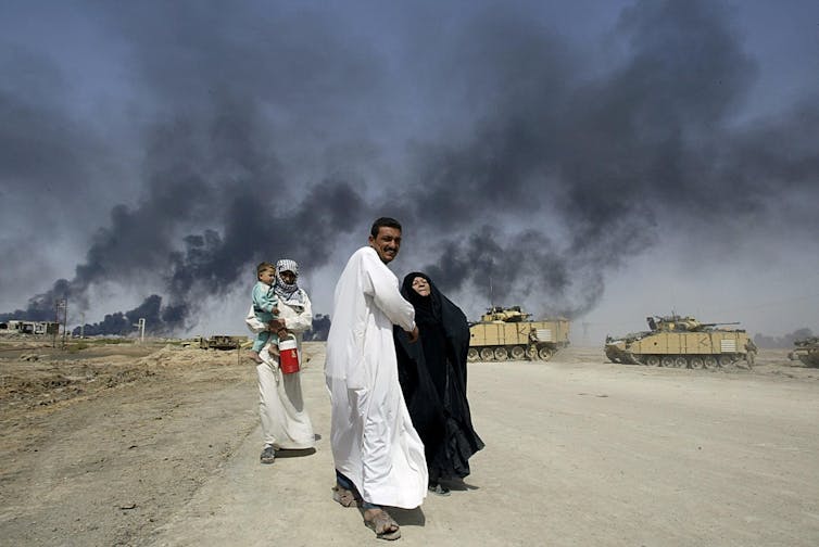 A man in white holds on to a woman covered in a black dress, while another man in white holds a baby and walks behind him. Smoke billows in the sky.