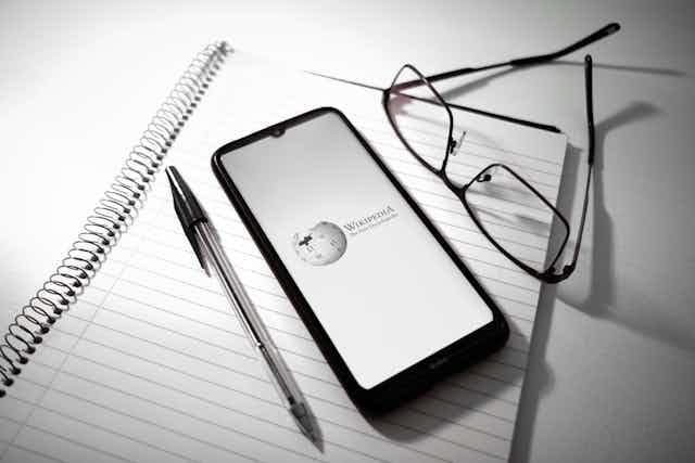 Wikipedia logo is displayed on a smartphone screen above a notebook next to a pen and glasses