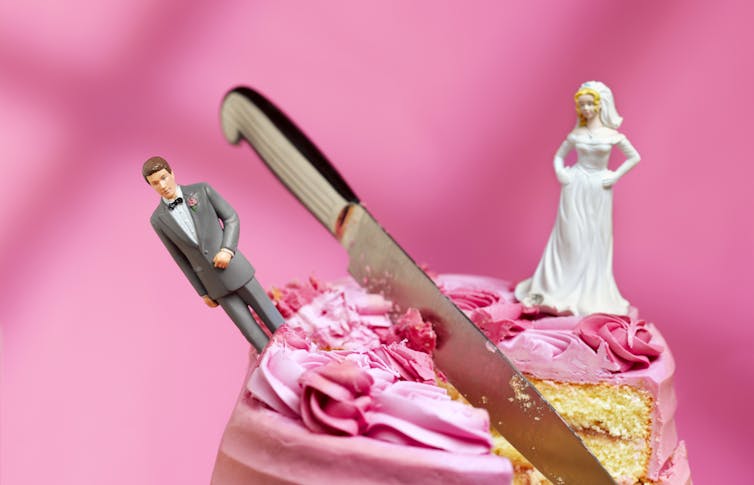 Bride and groom figurines arranged to look angry at each other on either side of a knife cutting a wedding cake in half.