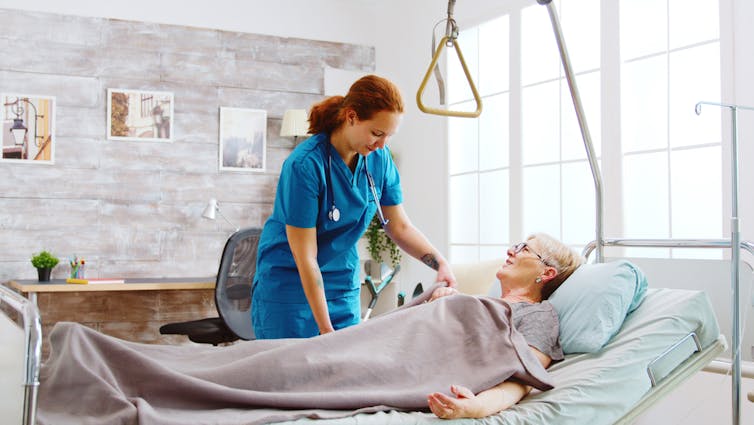 Une aide assiste une femme âgée installée dans un lit médicalisé posé dans son salon