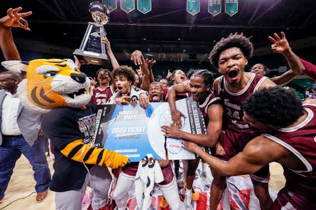 a bunch of excited basketball players wearing maroon jerseys and a person dressed up as a tiger hold a large piece of paper resembling a ticket as others hold up a trophy on a basketball court