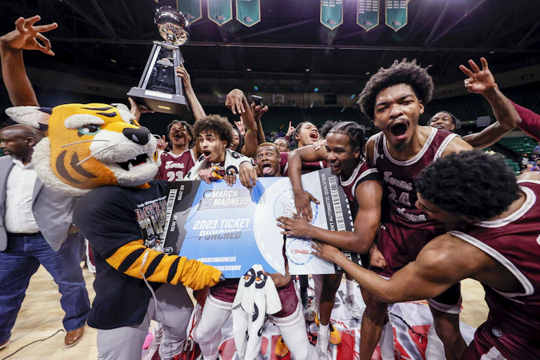 a bunch of excited basketball players wearing maroon jerseys and a person dressed up as a tiger hold a large piece of paper resembling a ticket as others hold up a trophy on a basketball court