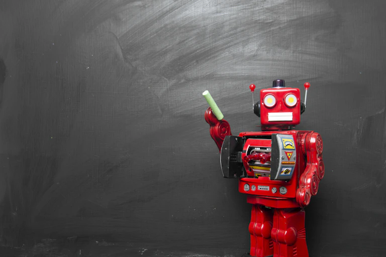 A toy robot holding a piece of chalk stands in front of a blank chalkboard