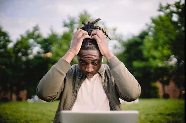 Man in park clutching head as reading laptop