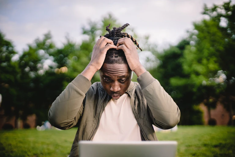 Man in park clutching head as reading laptop