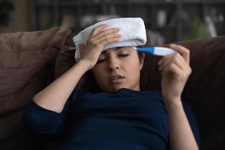 Woman on sofa with towel one forehead and thermometer in hand