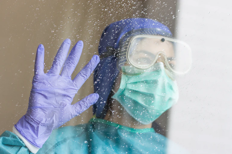 una sanitaria con gafas, mascarilla, guantes y el pelo tapado mira hacia la luz tras una ventana con gotas de lluvia.