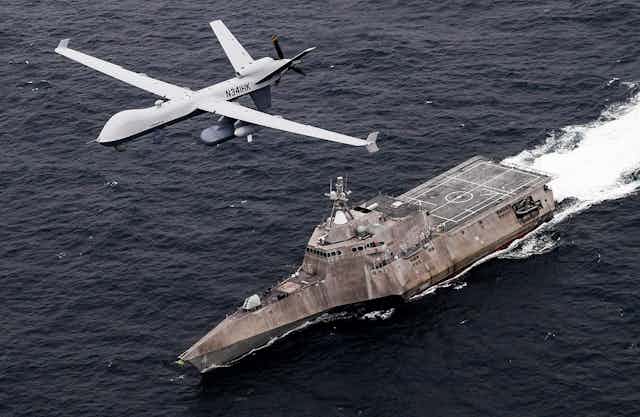 A small, unmanned aircraft hovers above a warship on the seas.