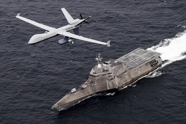 A small, unmanned aircraft hovers above a warship on the seas.