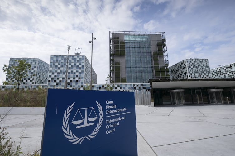 A blue sign says 'the International Criminal Court' outside of a large modern building structure.