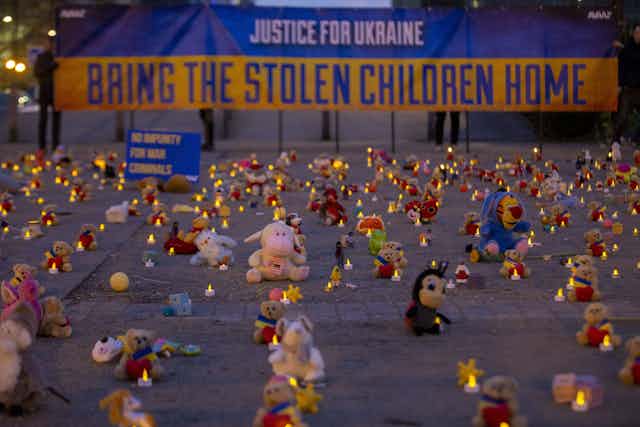 Stuffed animals are lined up in rows, with candles, in front of a big sign that says 'justice for Ukraine, bring the stolen children home.'