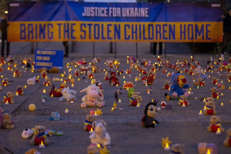 Stuffed animals are lined up in rows, with candles, in front of a big sign that says 'justice for Ukraine, bring the stolen children home.'