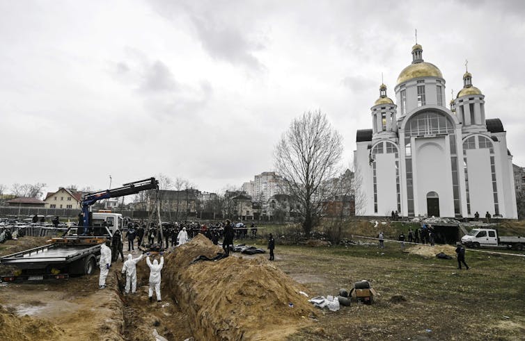 People in white appear to be digging in a large trench, outside of a white church.