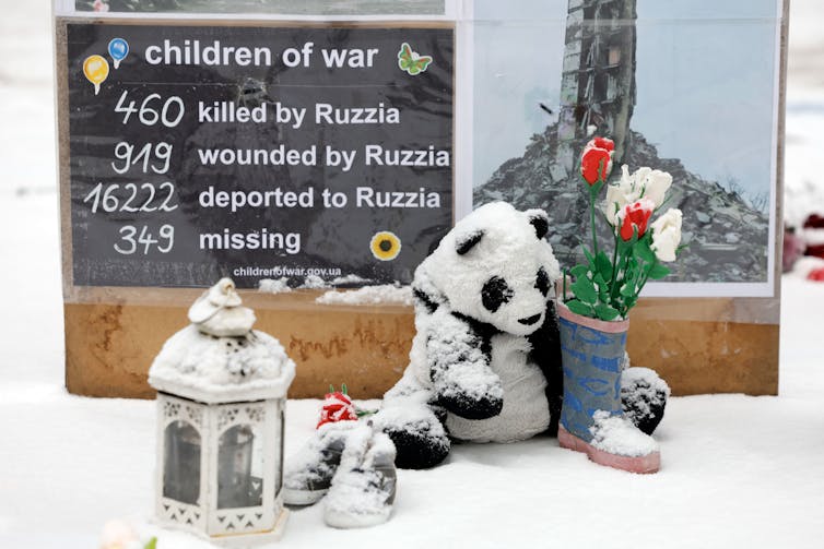 A stuffed panda bear is covered in snow and sits in front of a sign that says 'children of war. 460 killed by Russia, 919 wounded by Russia, 16222 deported to Russia, 349 missing.'