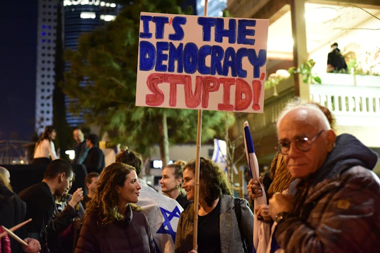 Israeli citizens demonstrating and holding up placards reading 'It's the democracy stupid'.