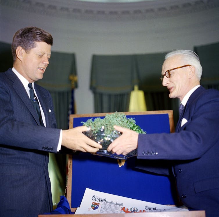 Two men wearing blue suits hold a green garland and look down at it.